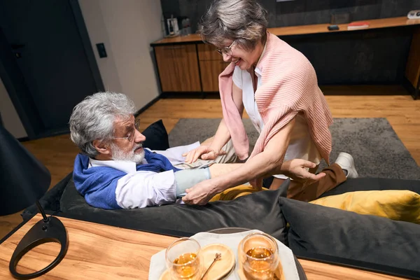 Optimistic wife measures the pressure of a gray-haired husband with a tonometer, the couple is located on a comfortable sofa